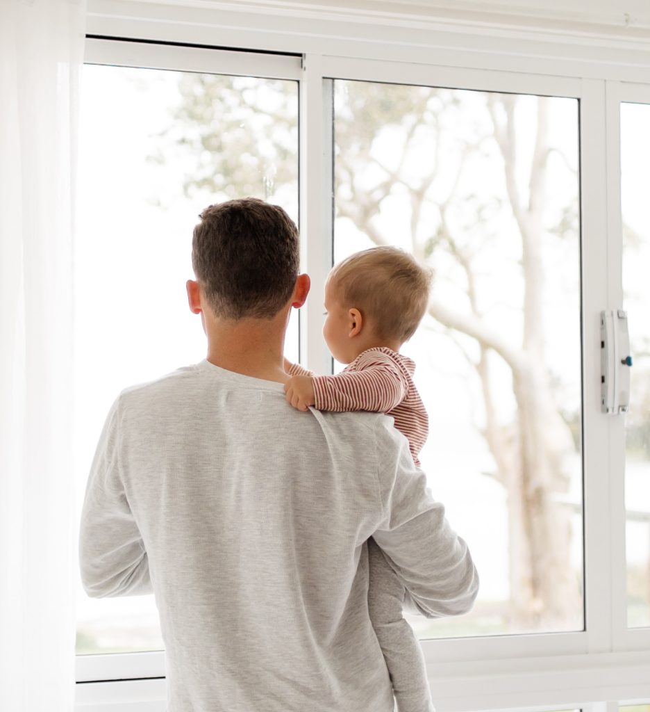 Isä ja vauva kuvattuna takaapäin. Isä pitää vauvaa sylissä ja he katsovat yhdessä ulos ikkunasta.