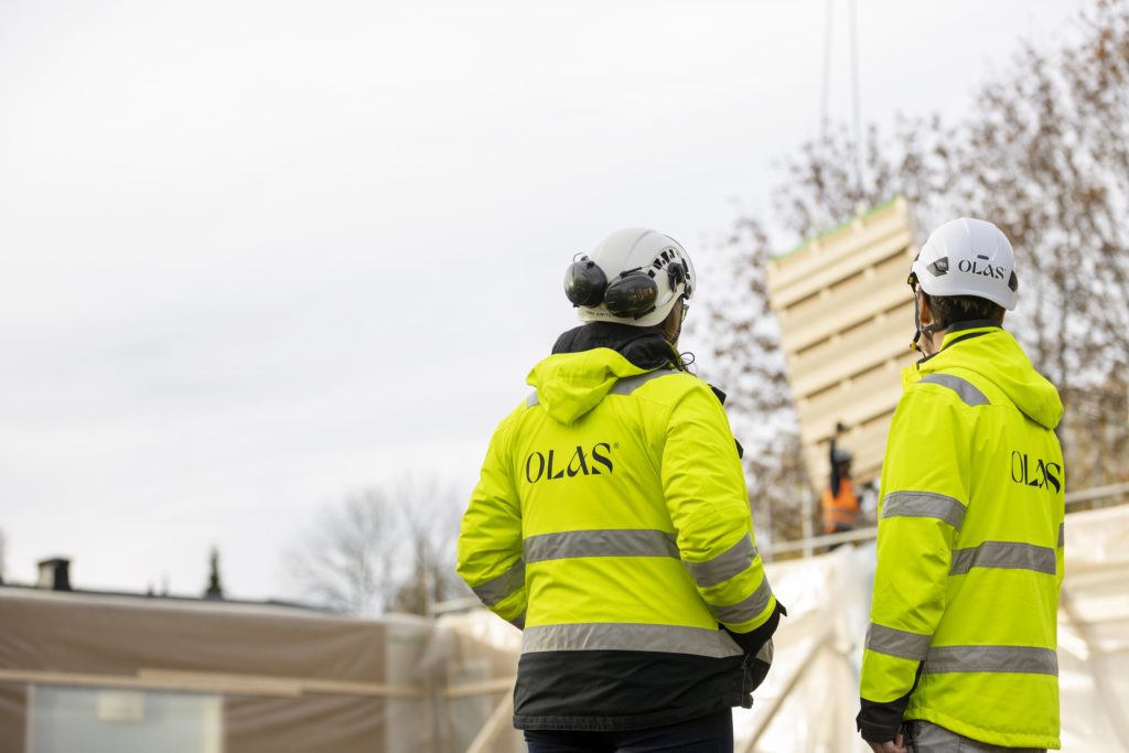 Kaksi rakennustyöntekijää keltaisilla Olas-työtakeilla rakennustyömaalla. Molemmilla valkoiset kypärät.