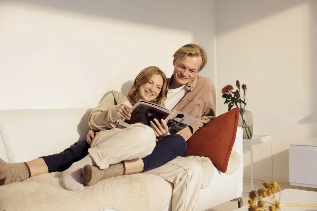Hymyilevät nainen ja mies makoilemassa sohvalla lukemassa lehtiä. Ilta-aurinko valaisee huonetta.